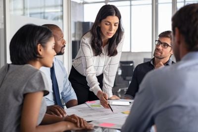 A woman leads a team discussion, pointing to documents as colleagues gather around a conference table.