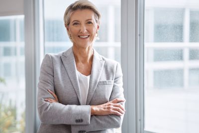 Smiling businesswoman in a gray blazer standing with her arms crossed in a bright office.
