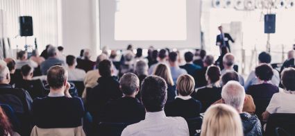 Conference attendees listening to a speaker presenting at the front of the room.
