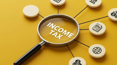 Magnifying glass highlighting “Income Tax” surrounded by financial icons on a yellow background.