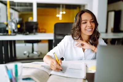  Woman wearing headphones and taking notes while looking at a laptop in an office.
