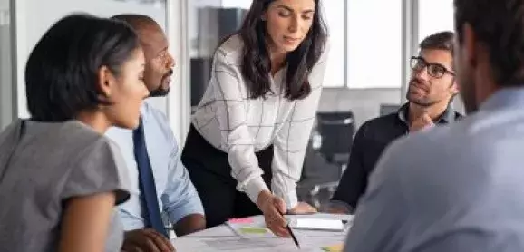 A woman leads a team discussion, pointing to documents as colleagues gather around a conference table.