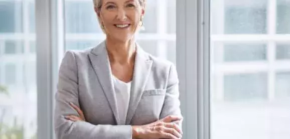 Smiling businesswoman in a gray blazer standing with her arms crossed in a bright office.