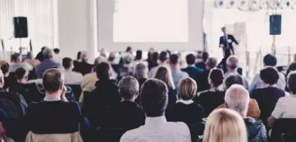 Conference attendees listening to a speaker presenting at the front of the room.