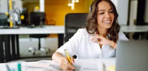  Woman wearing headphones and taking notes while looking at a laptop in an office.